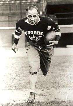 A football player in his mid thirties is seen running toward the camera with a ball tucked under his left arm.  He balding, and wearing a dark coloured uniform with the words "Crosse and Blackwell Chefs" on his chest.
