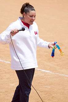 A standing brunette woman, wearing a white sweatshirt, holding a microphone in her right hand and a gold medal in her left.
