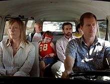 A film screenshot shows the family all seated in the Volkswagen Microbus as it is driven on a highway. The angle is from the windshield looking into the vehicle so that the majority of the interior can be seen. The mother is sitting in the front passenger seat with a dull expression on her face. The father is driving the vehicle with a smiliar expression. In the middle row, the daughter is looking down, listening to music from a CD player. Her uncle is seated on the right, looking to his left. In the back row, the grandfather is looking towards his grandson (who's face is slightly obscured by his father's head due to the angle). In the background, other vehicles can be seen driving on the highway.