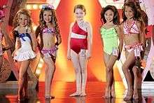 A film screenshot showing five girls standing on stage at the pageant at the end of the film. From left to right, the first girl (wearing a one-piece swimsuit) has her left leg placed in front of her right, the second girl (wearing a two-piece swimsuit) has her legs shifted in the opposite direction, at center the main character (wearing a one-piece swimsuit) is standing straight, the fourth girl (wearing a one-piece swimsuit) has her left leg placed in front of her right, and the last girl (wearing a two-piece swimsuit) also has her left foot forward. All of the girls are smiling towards different angles. The background of the stage is decorated and it is reflected on the shiny surface of the stage.