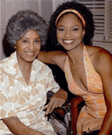 A dark-skinned woman with curly hair has her arm around an older woman in a wheelchair.
