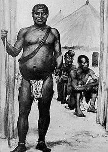 A black-and-white image depicting a middle-aged African king. Though at first glance a tall, strong-looking man, the subject's condition appears to have deteriorated with age; his belly appears severely bloated. He wears an animal-skin loincloth and a necklace made from what appear to be the teeth of various creatures. He holds a long spear in his right hand, which rests on the ground.