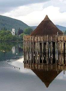 A small, brown conical structure sits on top of wooden piers set into a body of water. Ducks paddle through the water and in the near background there is a tree-lined shore with a white square tower showing amongst the trees. Tree covered hills and grey skies dominate the far background.
