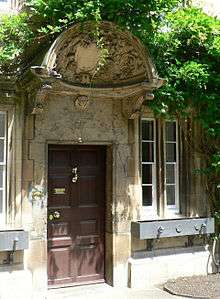 A large decorated shell-like stone canopy, with a shield in the centre and carved foliage around; beneath it, a wooden door with brass handle and knocker, set into a stone building; foliage grows above and around the canopy
