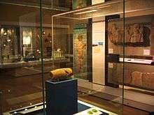 View of the Cyrus Cylinder in its display cabinet, situated behind glass on a display stand. Other ancient Persian artefacts can be seen lining the room in the background.