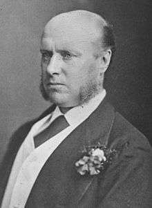 A balding man with long sideburns, wearing a suit with a buttonhole carnation