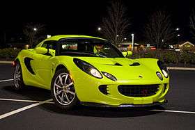 Picture of a 2005 Krypton Green Lotus Elise.  The car is positioned diagonally across parking space markings in an empty parking lot of a shopping mall in Ten Broeck, Kentucky, US. A number of stores are visible in the distant background. It is night and the car is illuminated by a tall overhead lighting post to the left front of the car (not visible within the framed picture) that casts a shadow almost directly under the car, but shifted slightly to the right rear. Numerous reflected lighting highlights are visible across the smoothly curved glossy body surfaces. The photo is taken from the right front of the vehicle approximately 8-10 feet away.  The front wheels are turned slightly to the left so that eight silver spokes and the center "Lotus" emblem is readily visible on the passenger side front wheel.  A red vertically positioned front tow hook protrudes from the black front egg-crate grillwork and casts a slight shadow down over the front plinth. The car has been modified with the addition of a front carbon fiber splitter.  The headlights and sidelights are lit.