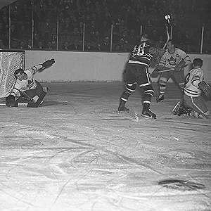 It is ice-level during action of a hockey game. A goal-tender reaches down with his blocker to stop a puck. An opposition forward and two of his own players are in front of his net.