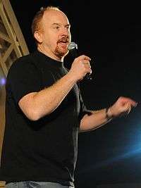 A balding man with red hair and a red goatee, wearing a black T-shirt, speaks into a microphone.