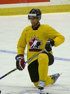 An ice hockey player wearing full equipment is kneeling on the ice with a smirk on his face. His uniform clearly indicates he is part of a national team, and his surname is stickered on the front of his helmet