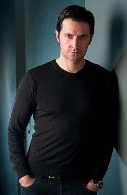 A serious looking dark haired man in his late thirties wearing a black t-shirt, and leaning against the wall