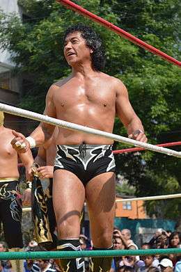 A color photograph of a Mexican male wearing black trunks with white markings, long black curly hair. He is standing in a wrestling ring at an outdoor event.