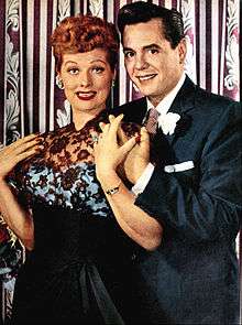 Colored glamorous shot of Lucille Ball and Arnaz standing. Both are smiling to the front. Ball at the left wears a ceremonial gown; Arnaz at right wears a tuxedo.