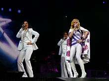 A blond woman, wearing a white leotard with purple stripes and white cape, singing on a stage. She is flanked by two female singer in white tuxedos. In the background the animation of a star can be seen.