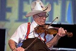 An elderlyl Luderin Darbone playing the fiddle.  He is sitting and wearing a cowboy hat.