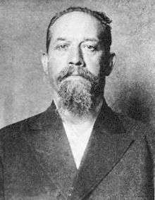 A black and white photograph of a man with short hair and goatee in a suit.