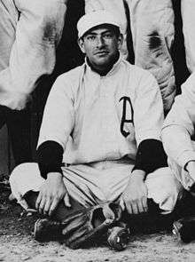 A man sitting on the ground wearing a white baseball uniform with an "A" on the chest has his baseball glove placed at his feet.