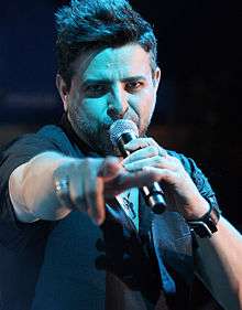 A man looking forward, with his right hand pointing forward and holding a microphone in his left hand.