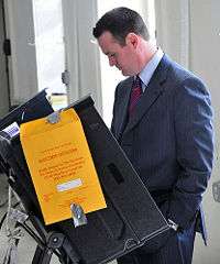 A young man with closely cropped hair and wearing a suit stands at an electronic voting machine