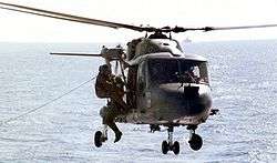 Two men in flight suits grapple on the side of a dark blue helicopter hovering over water.
