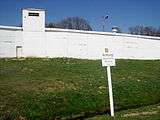 At the top of a grassy slope, there is a white concrete wall; treetops, a lamppost and the top of a guard tower are visible over the top of the wall. In the foreground, there is a white sign saying "Achtung! Bachmitte Grenze".