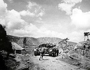 A tank advances up a hill followed by men in military uniform
