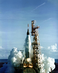 A rocket firing just above its launch pad, next to a metal structure, with a large cloud of smoke at its base and some smoke around the upper section of the rocket. Flame can be seen below the cloud.
