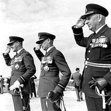 Three men in dark military uniforms with peaked caps and swords, saluting