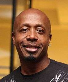 A image of a black man with an earring in his left ear. He is smiling and wearing a brown shirt.