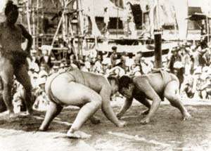 Two sumo wrestlers confront each other on a platform, their heads touching and their fists on the ground. To the side, a third man, also in a wrestling outfit, looks on. In the background, a crowd watches.