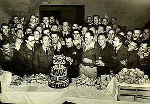 Large group of cheering men in military uniforms surround a man cutting a three-decker birthday cake on a long table