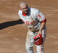 A man in a gray baseball jersey having just removed his red batting helmet at first base. His jersey reads "Phillies" in white and red script and has a number "8" on the left sleeve.