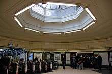 A station ticket hall with automatic ticket gates on the left and passengers waiting to buy tickets from a machine in the wall. A large octagonal roof light occupies the central portion of the ceiling with a deep convex moulding around the opening.