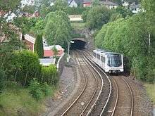 A double-track train line down the center of the image after emerging from a tunnel. The line is surrounded by houses and trees, and a white three-car train is halfway down the line on the right side.