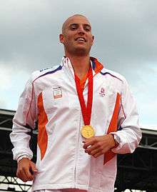 Shaven headed man with a gold medal hung around his neck on a red ribbon, wearing a watch on his left hand and a white tracksuit with small pieces of blue and orange.