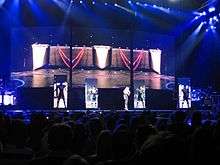 A stage with four rectangular screens which display a black geometric patterned screen approaching. In front of the screen stands Madonna and her three dancers, clad in a robotic outfit