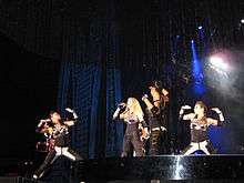 Left profile of a blond female in a tight black dress with gloves and wrist bands singing. A number of similarly dressed dancers encircle her while making gesture with their hand. Blue light is visible behind her and a metallic cover is present above their head.
