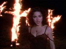 Madonna in front of a pair of burning crosses. The woman is wearing a dark, brown dress and looks towards the camera