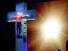 Madonna standing on a blue crystal cross, wearing a red shirt and dark brown pants. Her hands are spread apart along the cross's breadth as if she has been crucified. Behind the cross, a backdrop is centrally illuminated