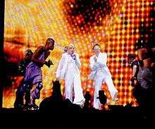 A female blond performer onstage wearing a white tuxedo and singing to a microphone in her left hand. A similarly dressed woman claps on her left. They are encircled by a group of guys in roller skates. The backdrop display disco lights in red and yellow.