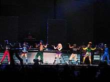 A long line of people in colorful clothes striking a pose on a stage with both their hands pointed up but their palms straight and looking to the right. Middle of them is a blond woman who sings into a microphone in her left hand.