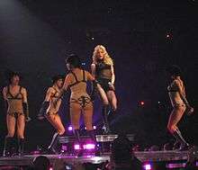 A blond female stands on a raised platform of a stage. Her wavy hair falls around her and she wears a black top and high boots while holding a microphone in her left hand. She is encircled by female dancers in short black hair and nude body suits which are lined in black. They appear to be hopping.