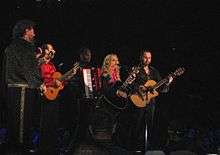 A blond woman sits on a long bar stool in front of a microphone playing a black acoustic guitar. The woman's hair is wavy and she wears a black dress with a number of pink threads around her neck. On her right two middle aged men playing acoustic guitar are visible.