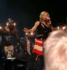 A blond woman on stage. She wears red-and-white striped skirt and is wearing a black top with zippers. She's singing into a microphone she holds with her arm. To her right there is a dancer wearing a mask and wearing black clothes. The head of a blonde audience member can be seen.