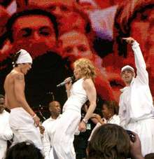 Left profile of a blond female who is singing into a microphone in her right hand. She is wearing white trousers and white, sleeveless overcoat with bangles in her hands. The woman is flanked by two male dancers and behind them another group of people is seen, all dressed in white garments. The backdrop displays blurry faces of the crowd.