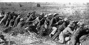 Boer War scene. Men of all ages wearing hats and bandoleers crouch in a line, rifles pointed