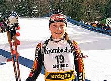 A young woman in multicoloured winter sportswear and with the number 19 on her jersey smiles into the camera, standing in a field covered with snow. A forest and many onlookers can be seen in the background. She holds ski poles in her right hand and has a rifle on her back.