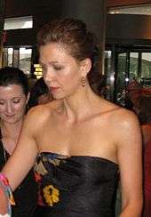 A brown haired woman looking away from the camera. Her hair is tied back, and she is wearing gold earrings and a shoulderless, sleeveless black dress with a yellow, red, and blue pattern