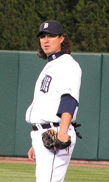 A man wearing a white baseball uniform with a navy blue "D" on the chest, a navy blue cap bearing a white "D", and a baseball glove stands on a baseball field