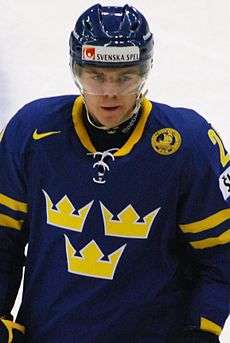 An ice hockey player standing, facing towards the camera. He is wearing a blue helmet and a blue and yellow uniform.